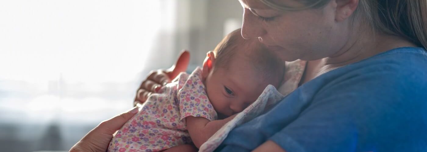 Young woman sitting on bed holding newborn daughter